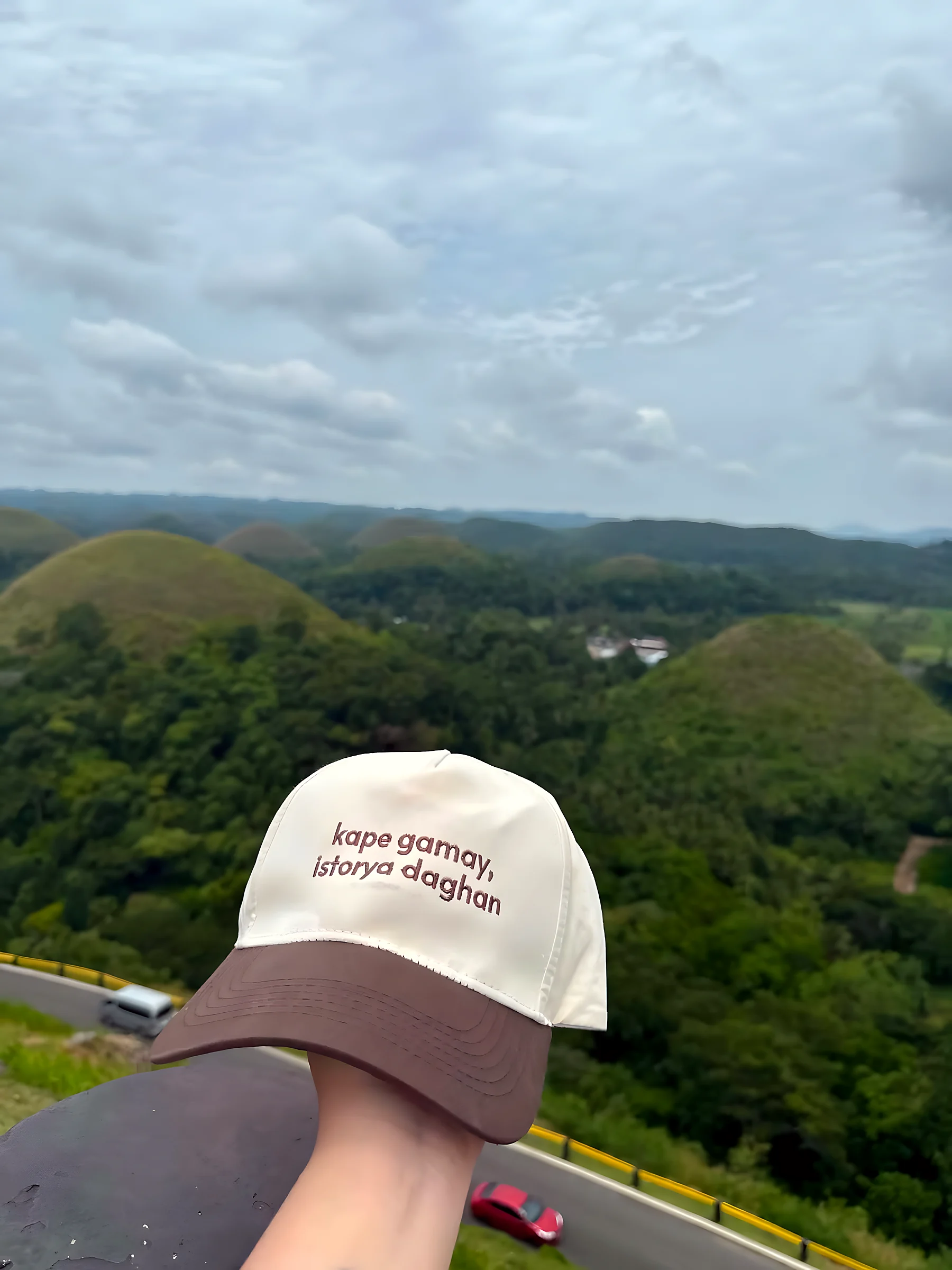 A cream cap with a brown brim, printed kape gamay, istorya daghan, held in front of green hills under a cloudy sky.