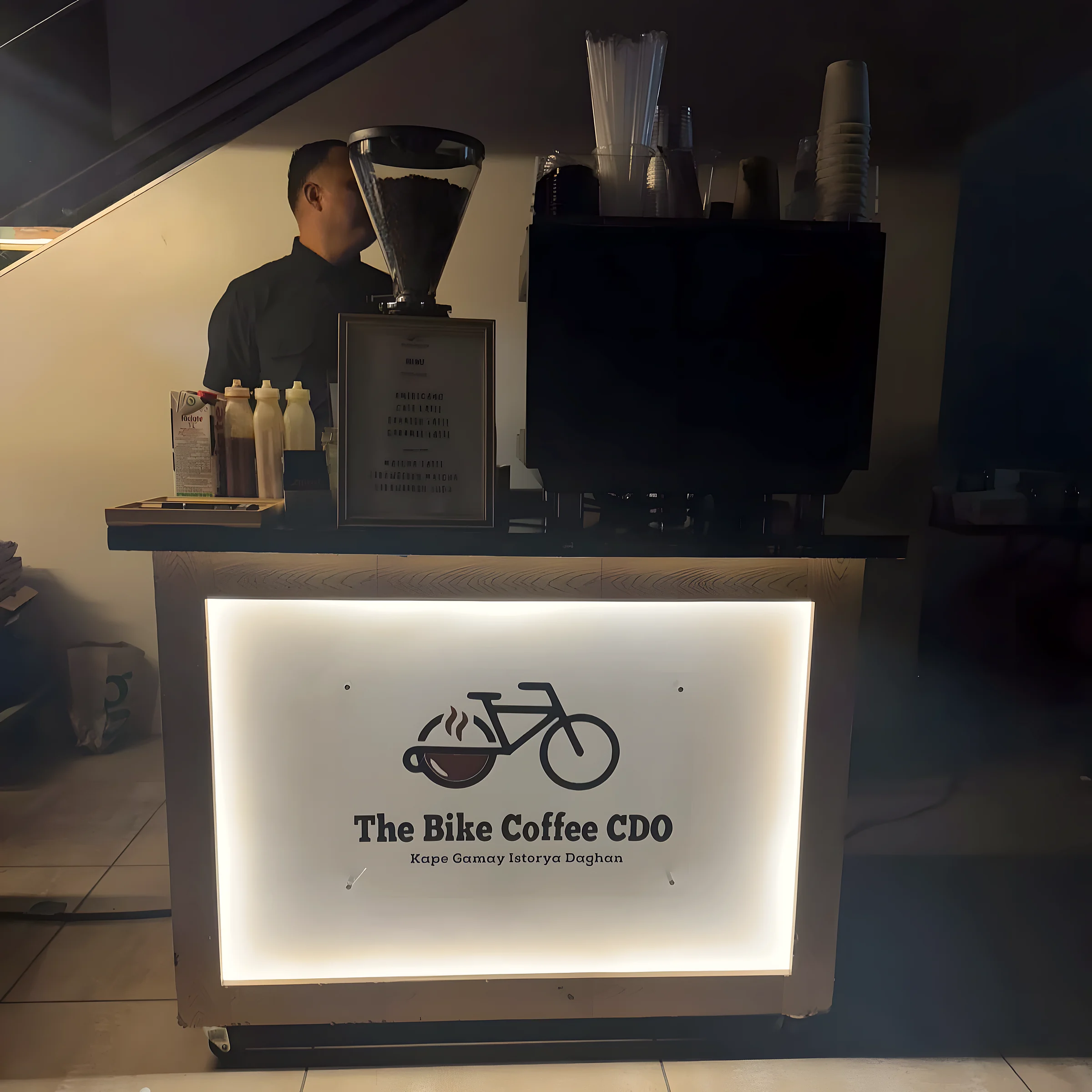 A coffee cart in a dim room. Its front panel is lit from inside and shows the bike-and-cup logo and the name The Bike Coffee CDO. A barista works behind it with a grinder and an espresso machine on top.