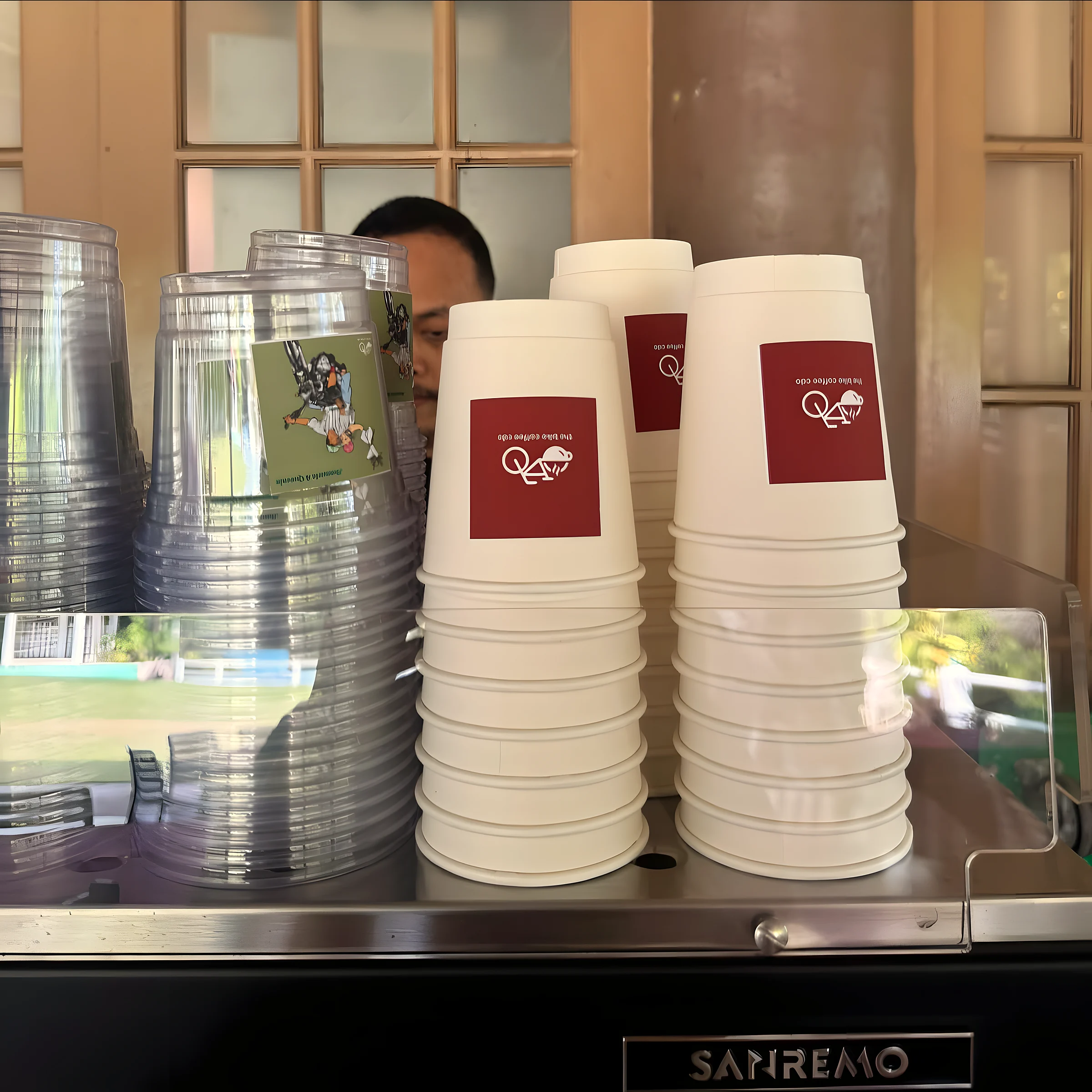 Stacks of white cups with maroon logo labels and clear cups, on a bar beside an espresso machine.