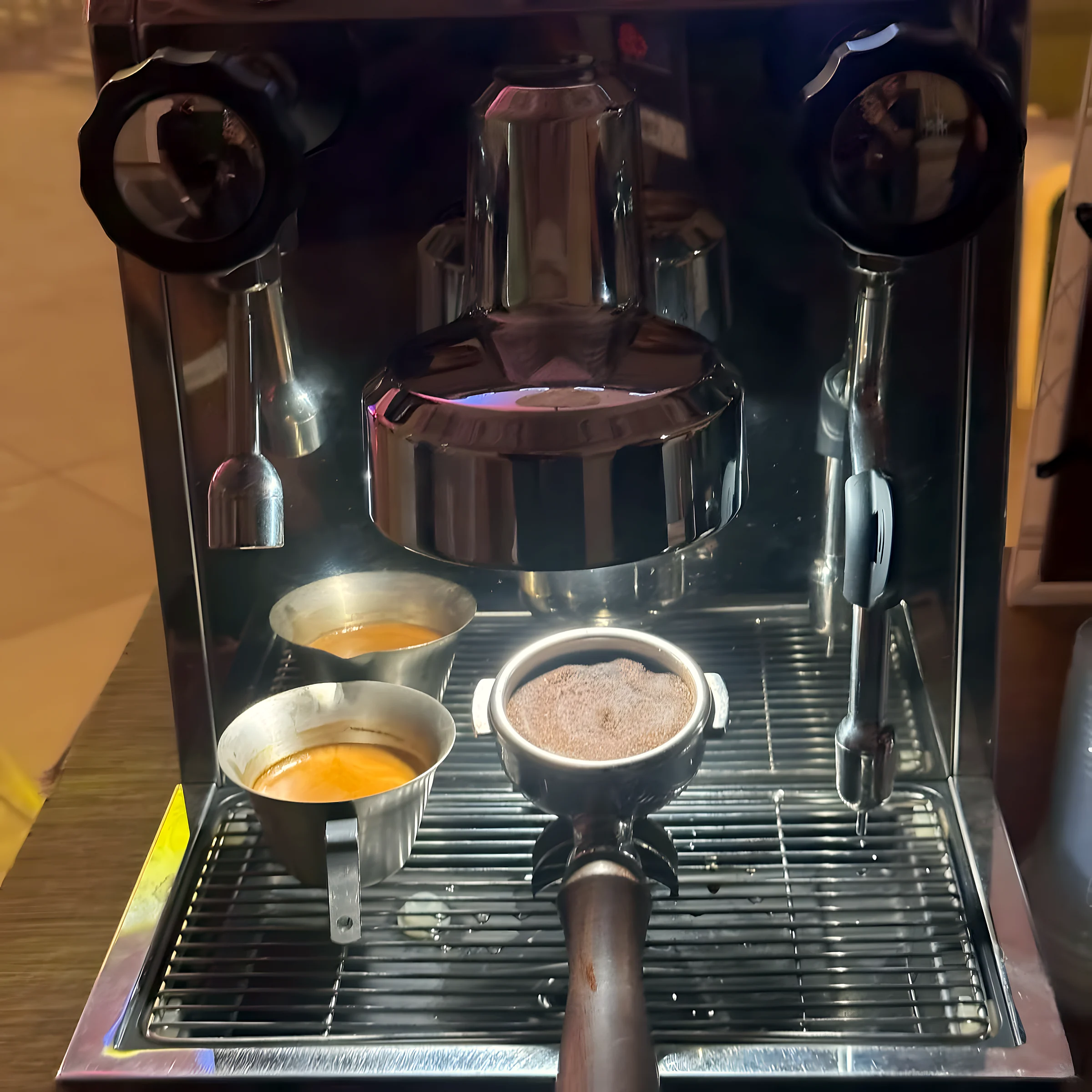 A steel espresso machine with a portafilter locked in and two shots pulled into small metal cups on the drip tray.