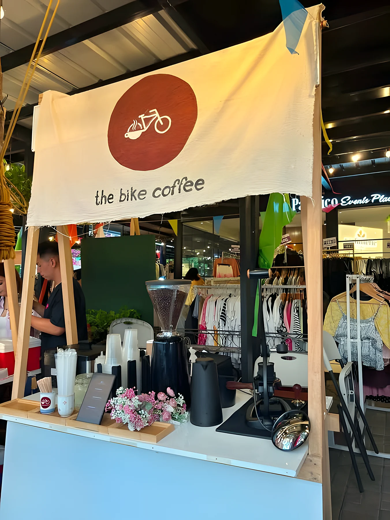 The pop-up stall at a festival, a brown disc logo on the banner, a barista behind a counter with a grinder and flowers.