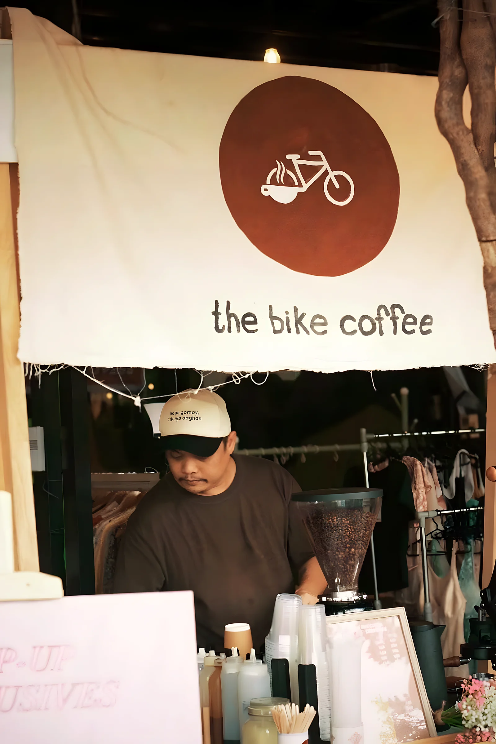 A white canvas banner with a brown disc and the bike logo hangs over a pop-up stall. A barista in a cap works a grinder below it.